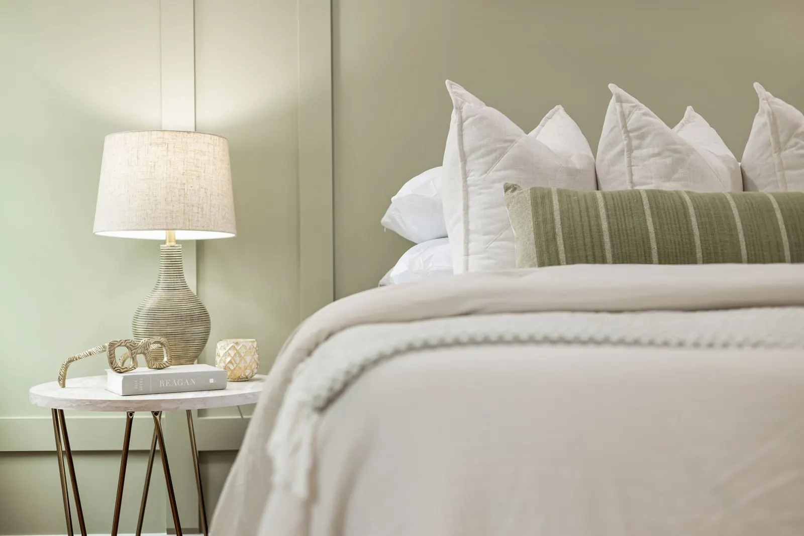 Bed with white bedding and a striped green lumbar pillow beside a table lamp on a round side table