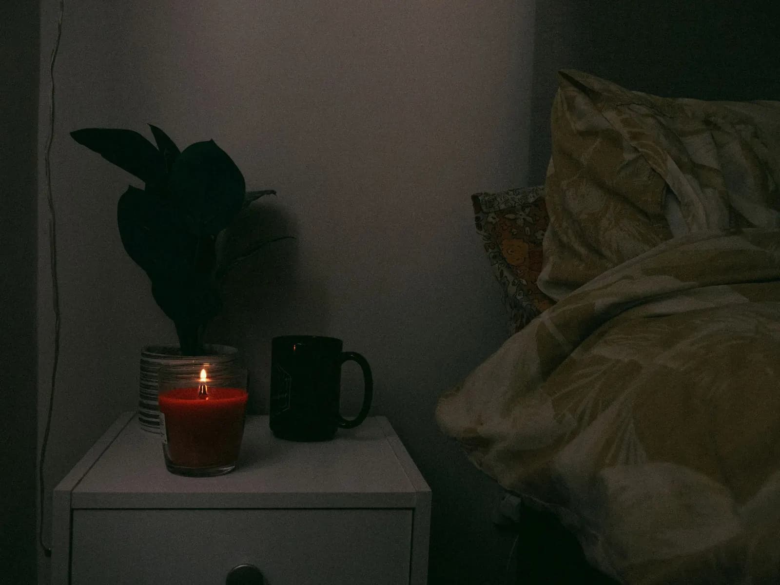 A cozy nightstand scene with a candle, mug, and plant beside a bed, creating a warm atmosphere.