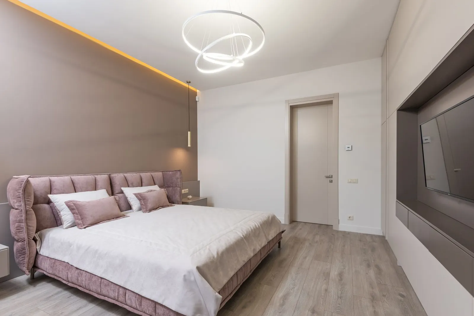 Interior design of comfortable bedroom with light brown and white painted walls furnished with bed with pink upholstery decorated with pillows Illuminated by