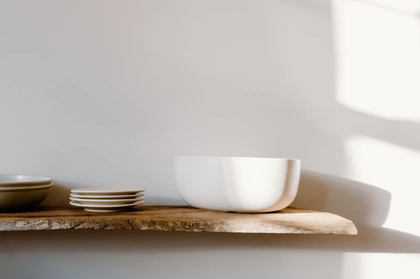 Elegant white ceramic bowls and plates on a rustic wooden shelf, showcasing minimalist design in natural light.