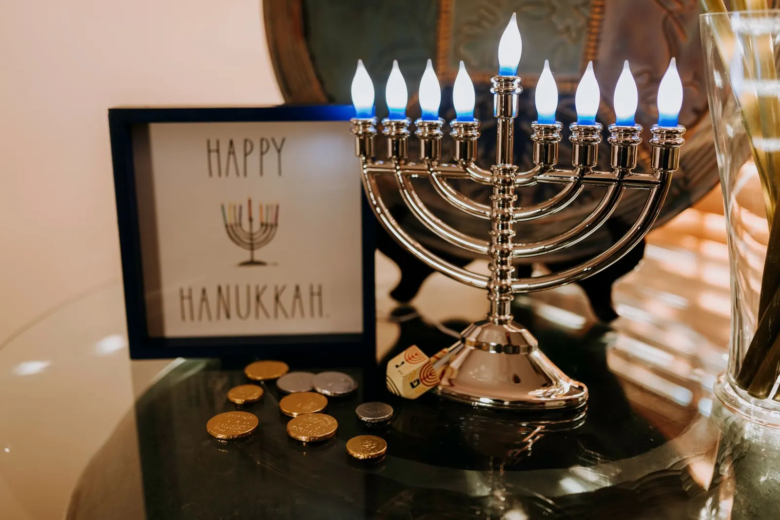 Silver menorah on a metal tray beside blue and white candles, dreidels and gold coins on a white table