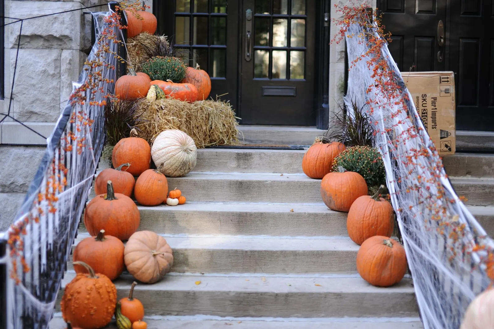 Stone steps lined with uncarved orange and white pumpkins, mums and hay bales below a black front door