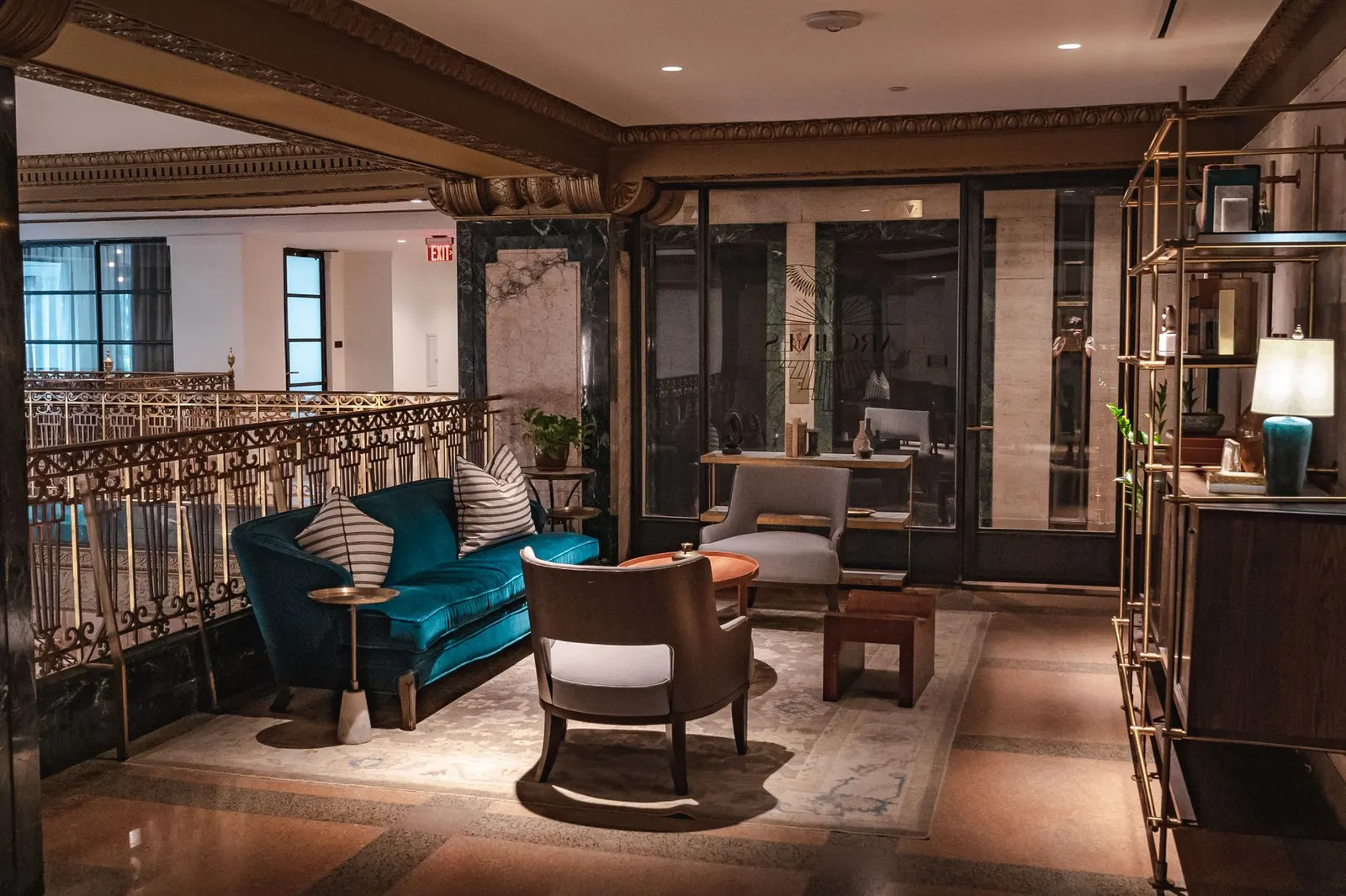 Lounge with a teal velvet sofa, curved wood armchair, gilded iron railing and brass shelving with a lamp