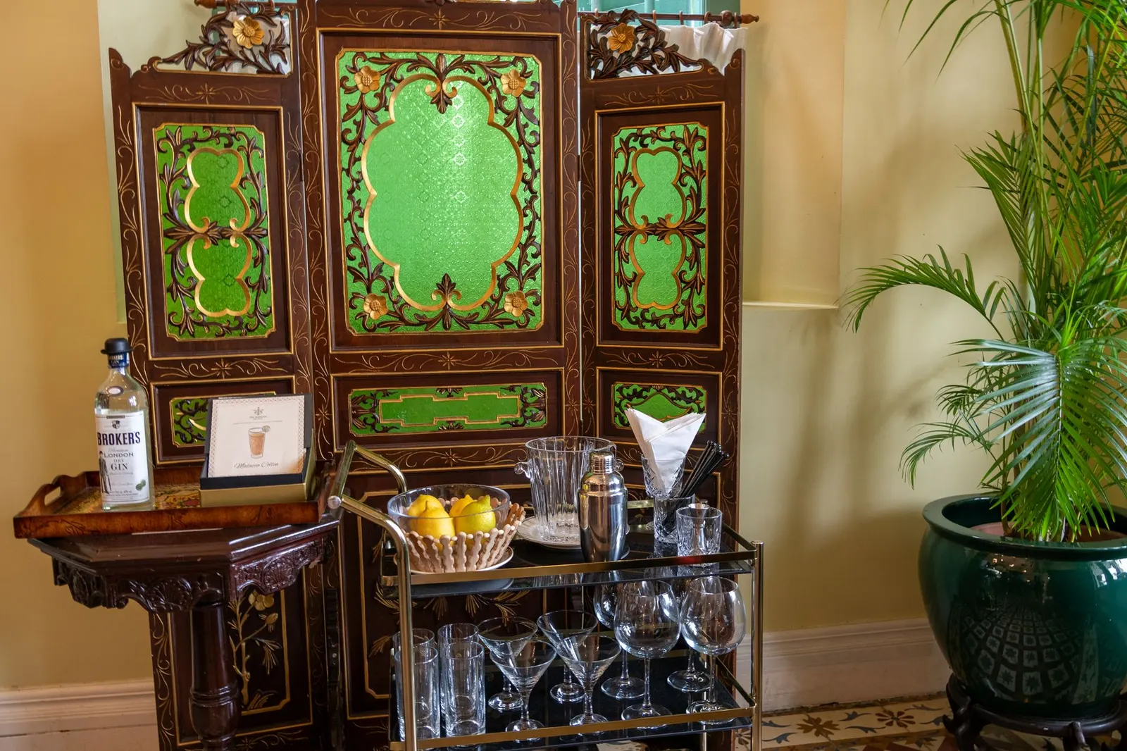 Bar cart with glasses, a bottle and a bowl of lemons in front of an ornate carved green cabinet