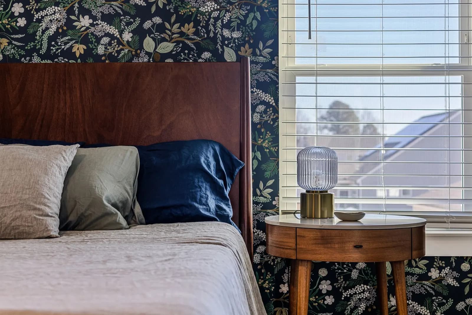 Elegant bedroom featuring floral wallpaper, stylish pillows, and natural light from a window near a nightstand.