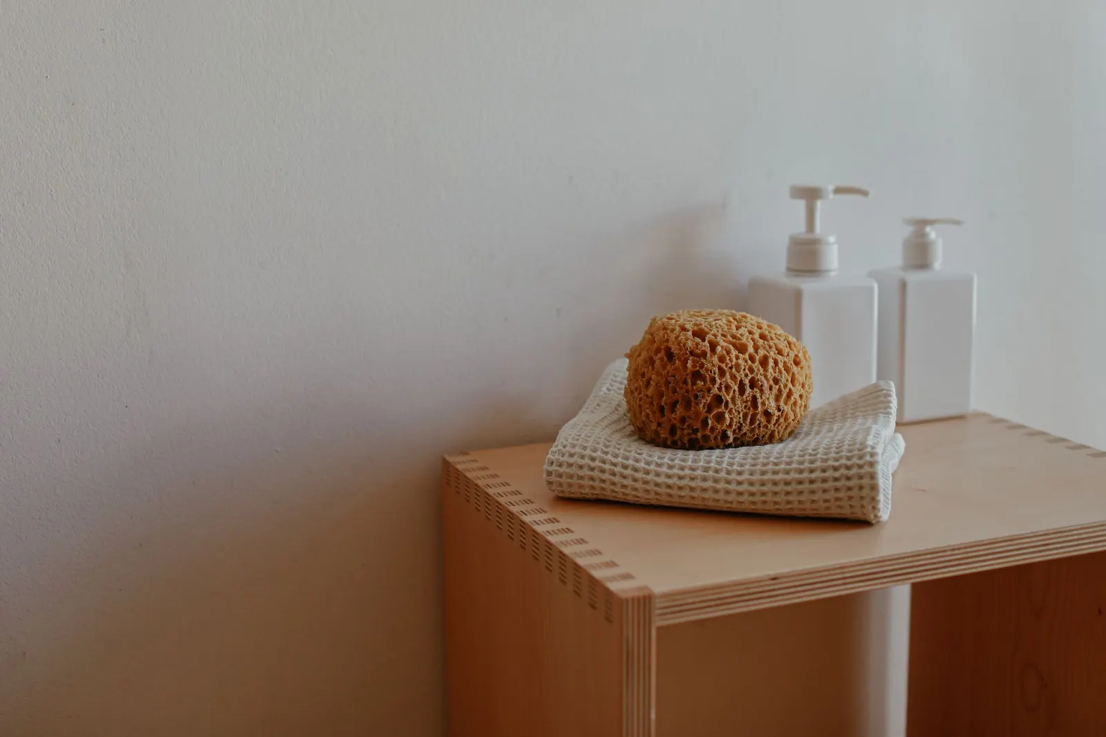 Close-up of a bathroom shelf featuring a natural sponge, towels, and pump bottles.